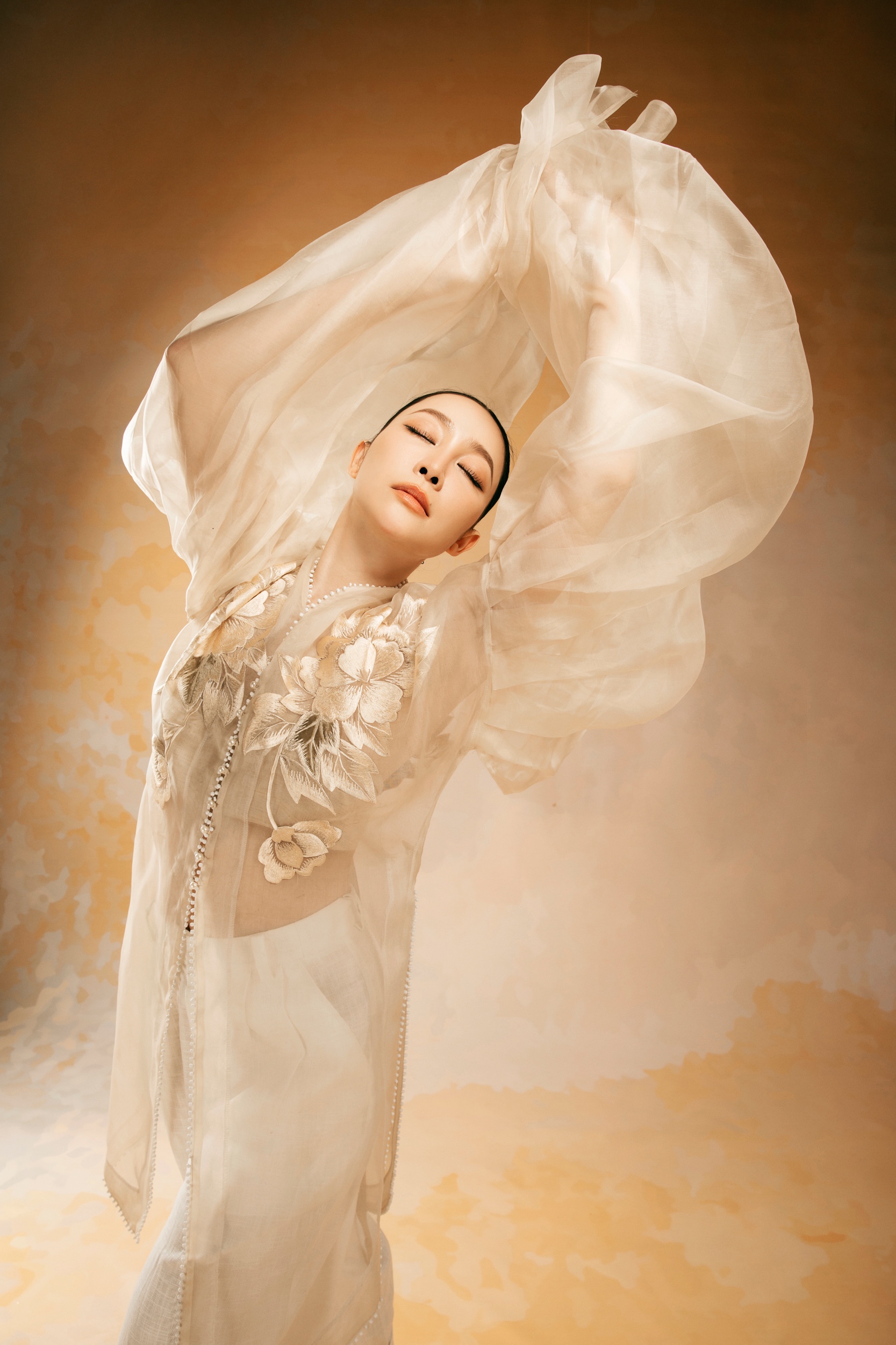 Linh Nga in a sheer ivory embroidered ao dai, arms raised with flowing fabric and eyes closed.
