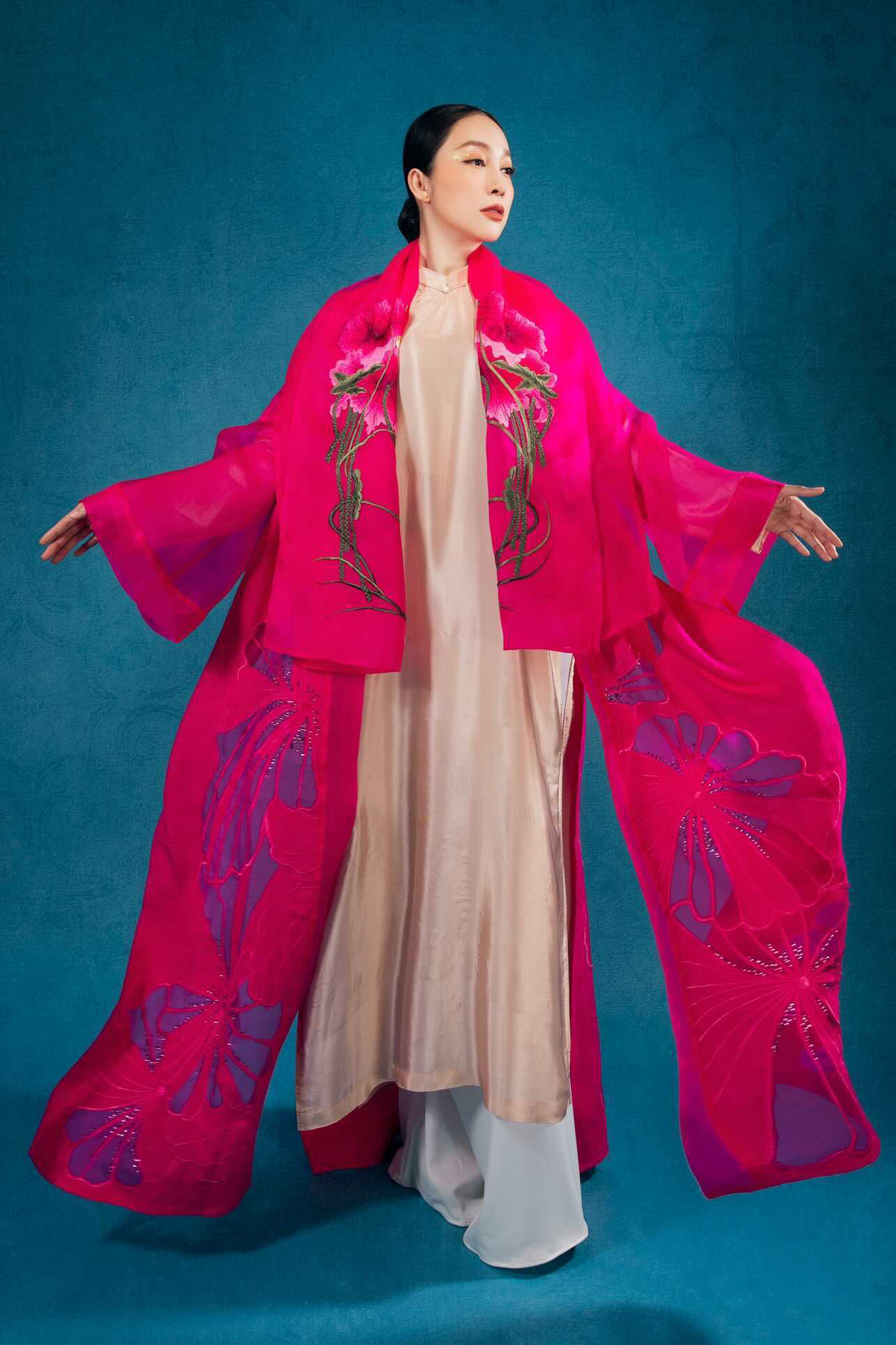 Linh Nga in a magenta silk robe with cut-out floral panels over a cream ao dai, arms outstretched, against a teal backdrop