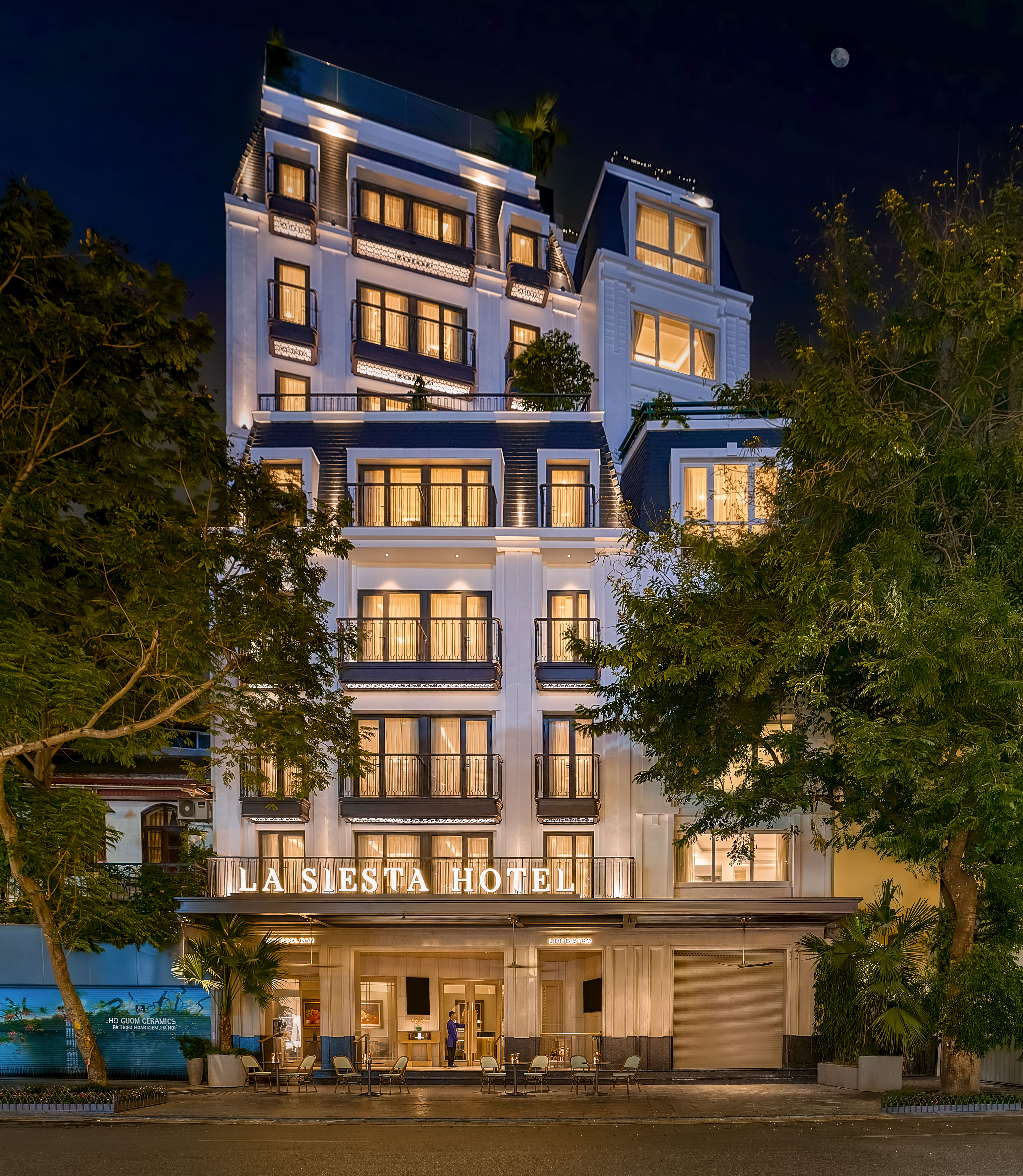 The white colonial-style facade of La Siesta Hotel on a tree-lined street in Hanoi's Old Quarter by day