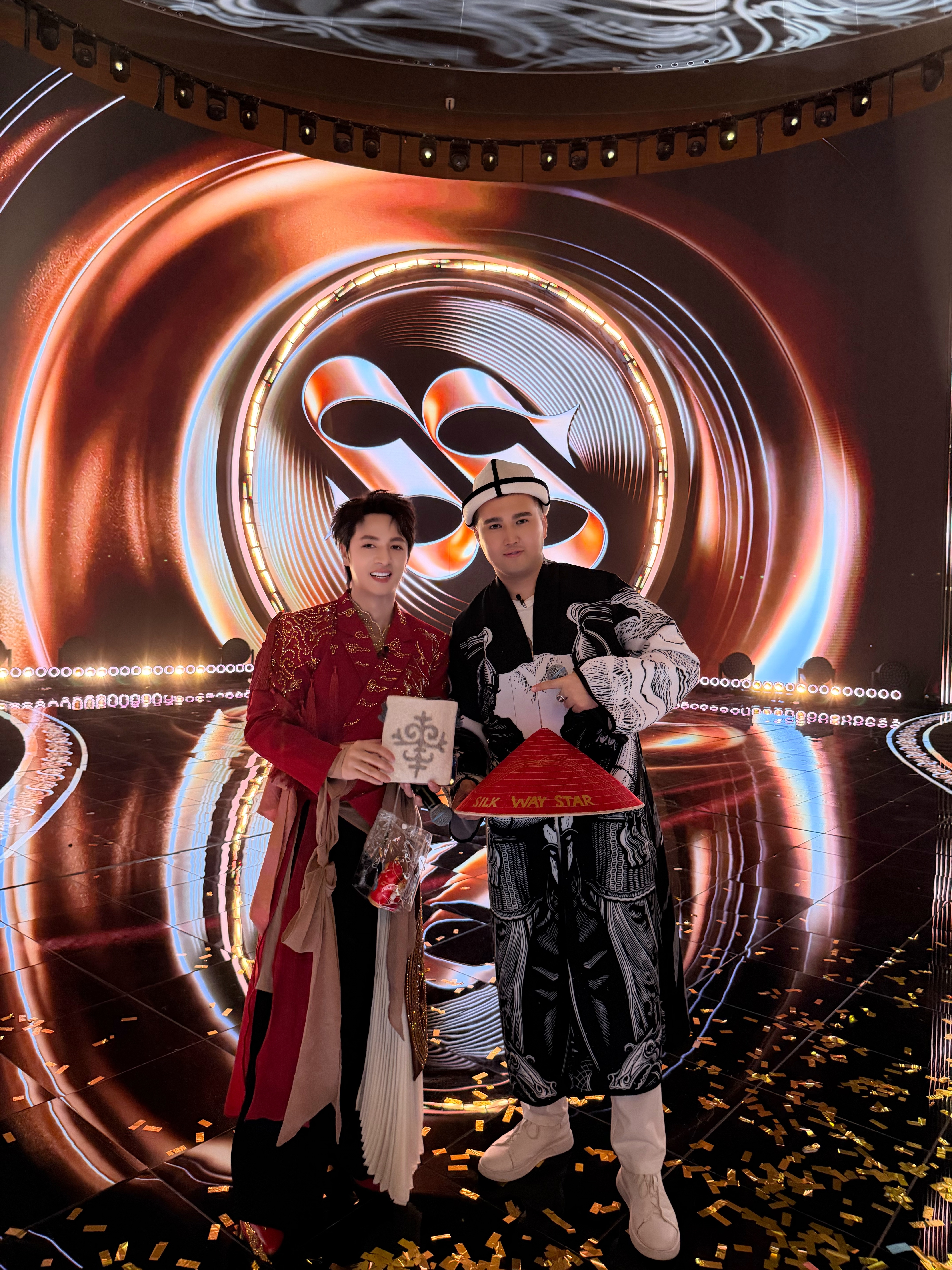 Đăng Khôi in red stands with a fellow contestant holding a "Silk Way Star" conical hat amid falling confetti.