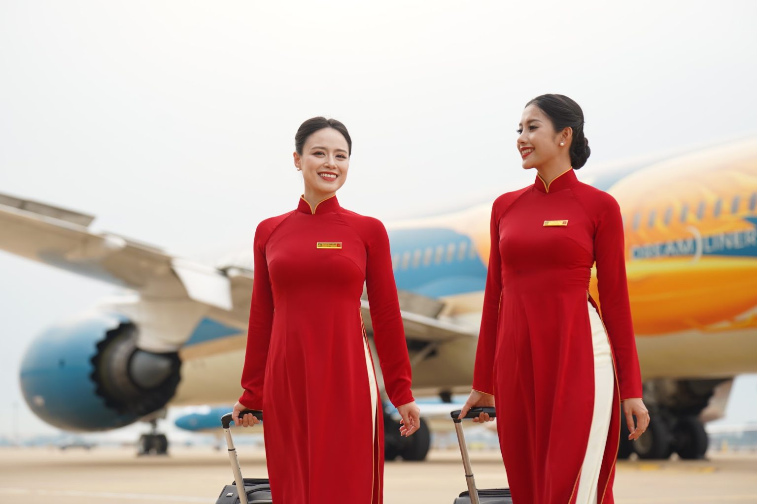 Two Vietnam Airlines flight attendants in red áo dài uniforms beside a Boeing 787