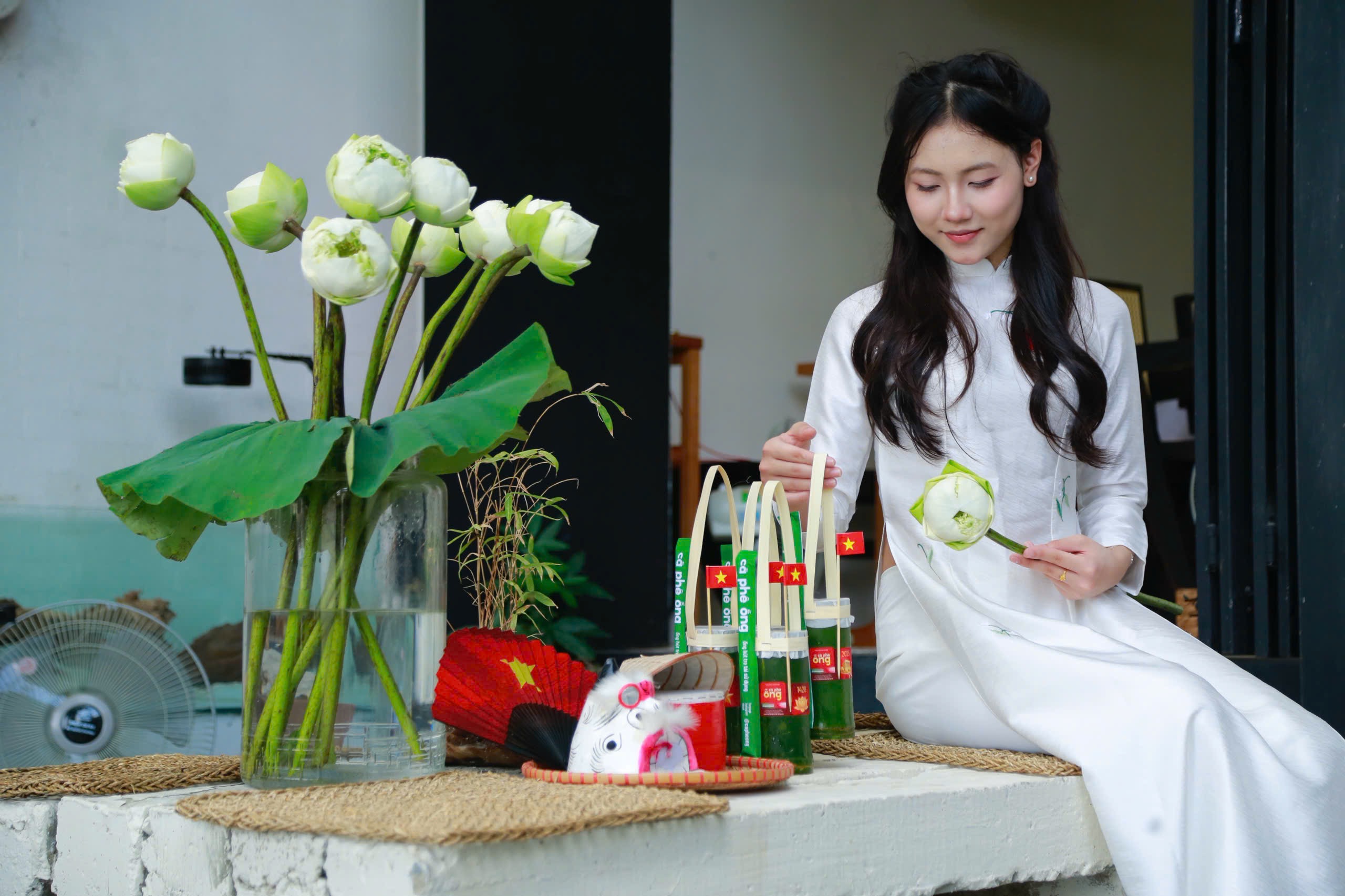 A young woman in a white áo dài holds a lotus bud beside special-edition cà phê ống bamboo tubes