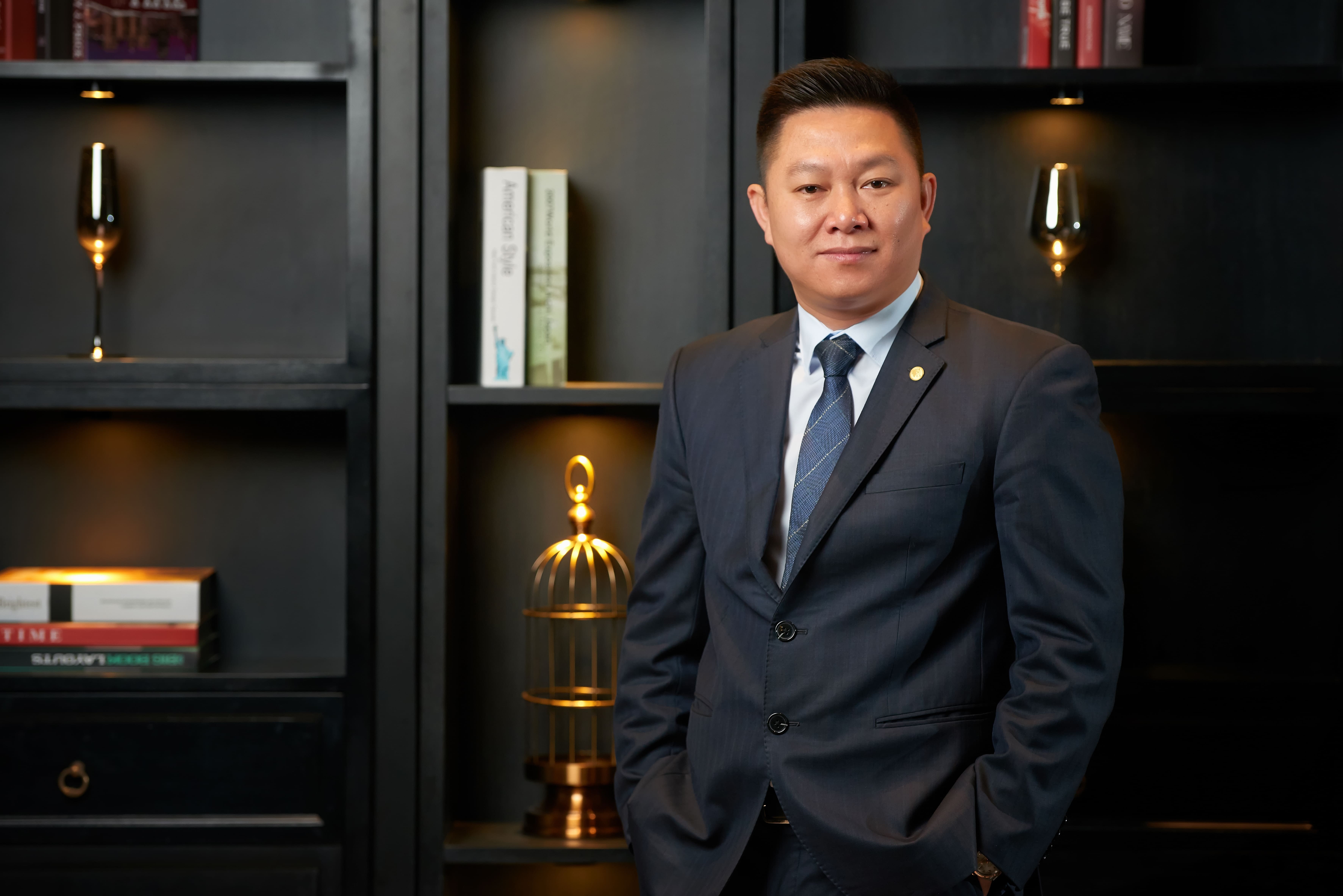 Do Van Dan in a dark suit, standing before a black bookshelf