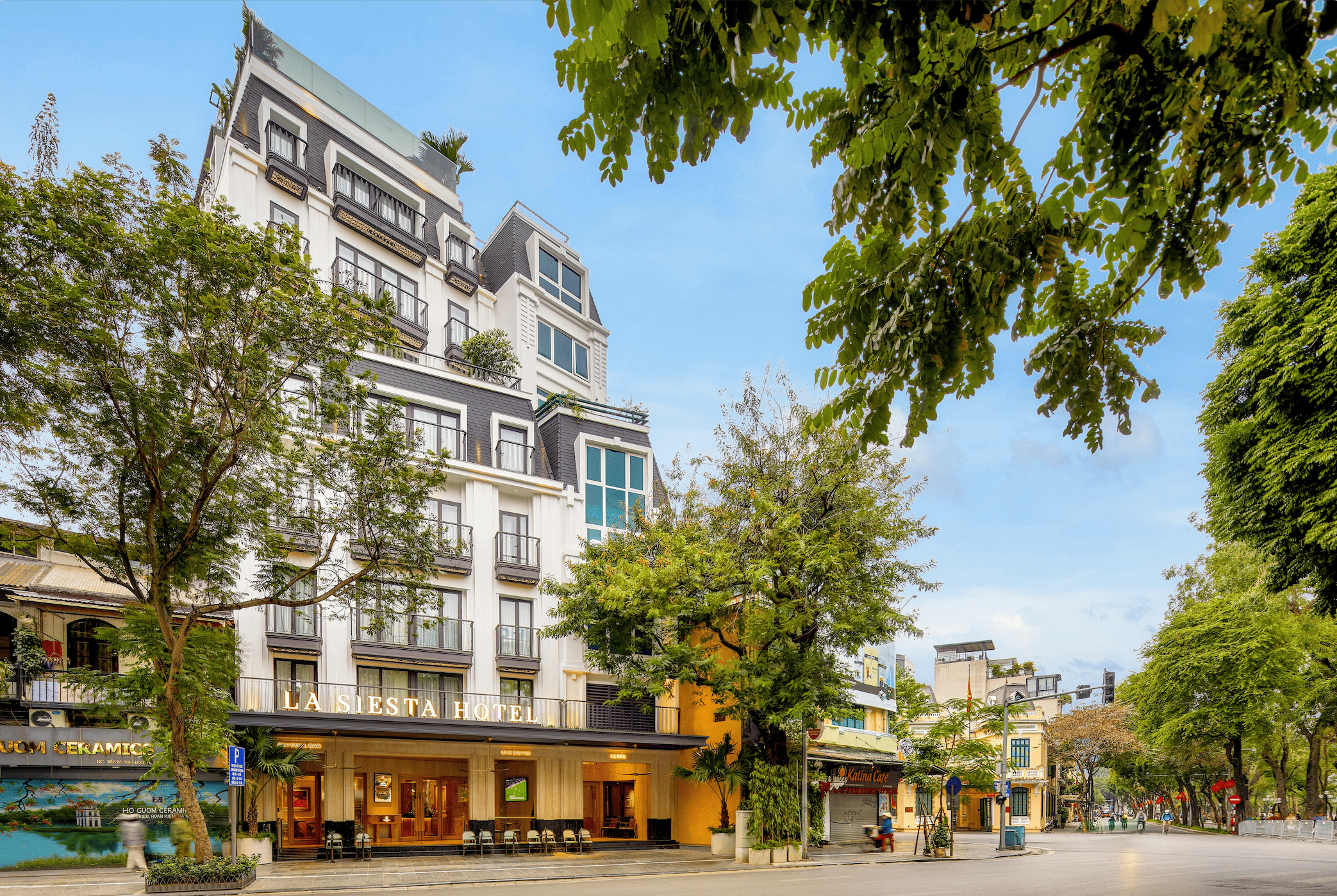 The white colonial-style facade of La Siesta Hotel on a tree-lined street in Hanoi's Old Quarter by day