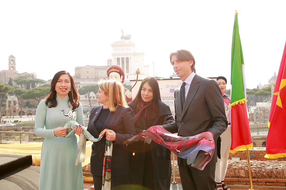 Officials and the designer holding Vietnamese silk at an event in Rome, with the Altare della Patria and Italian and Vietnamese flags behind