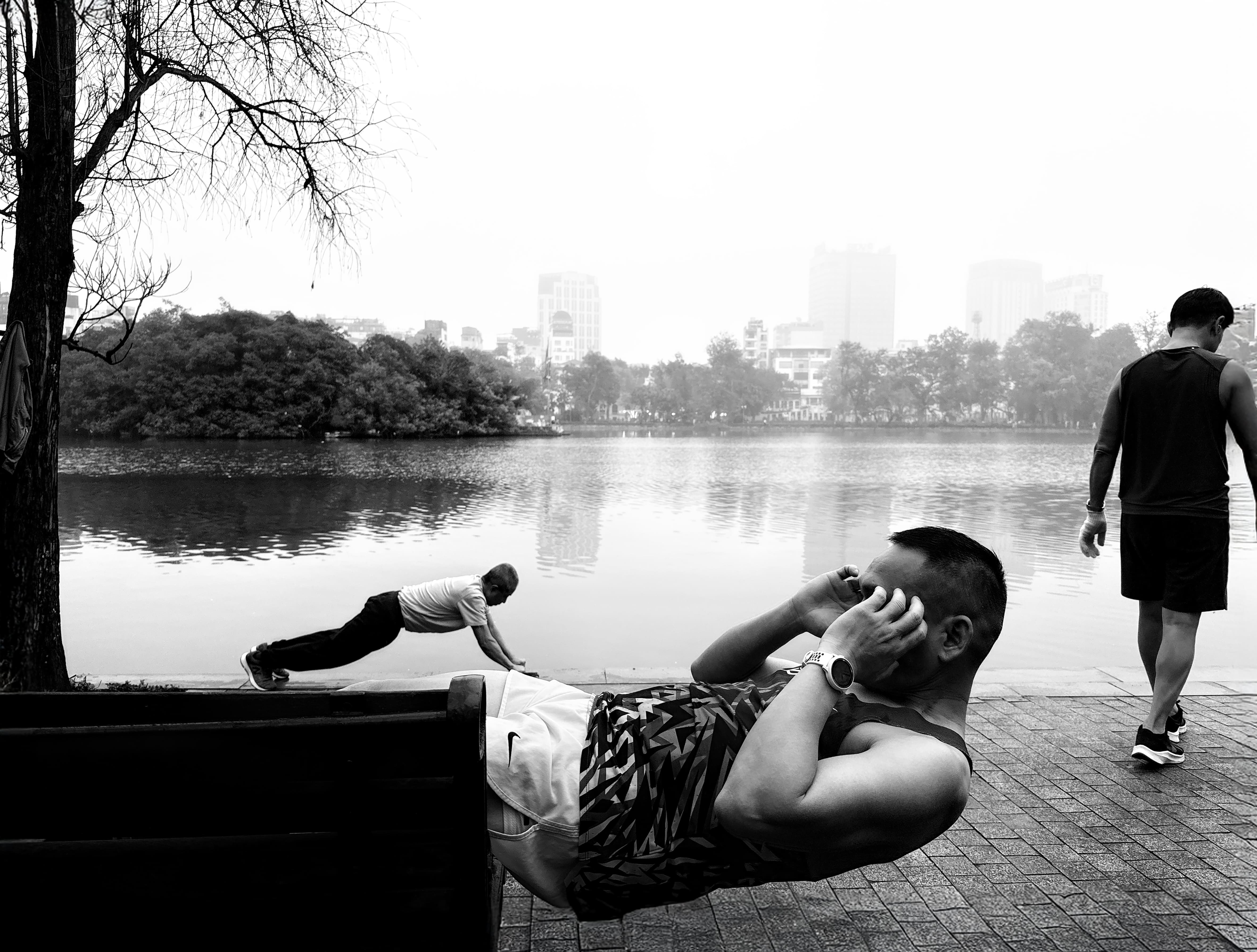 A black-and-white street scene by Hoan Kiem Lake, a man doing sit-ups on a bench while another exercises and a walker passes
