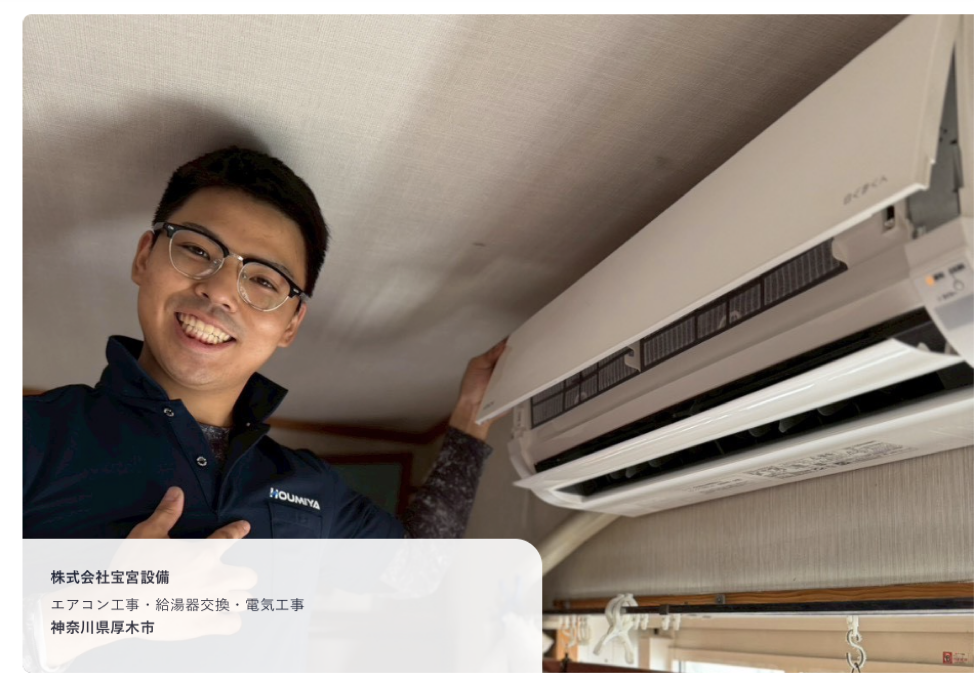 A smiling technician in glasses and a navy work jacket reaches up to a wall-mounted air conditioner.