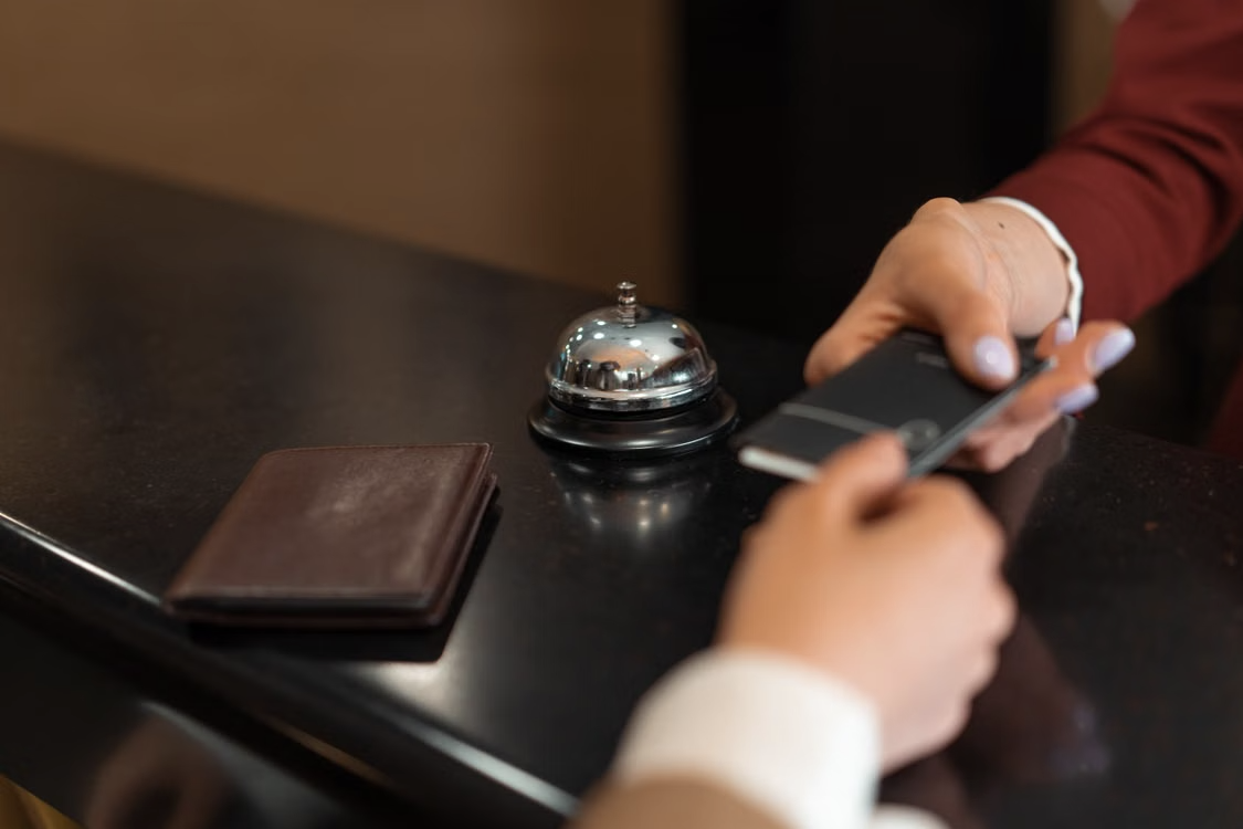 A hand passes a dark card across a black hotel reception desk, past a silver service bell and a brown leather wallet.