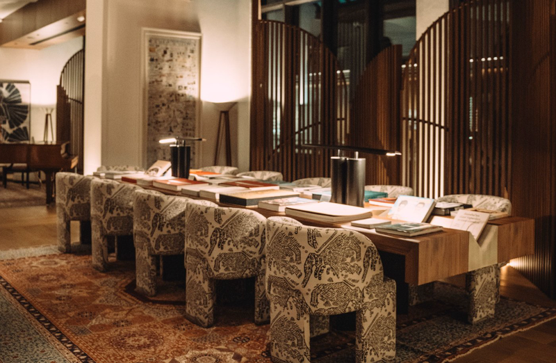 A hotel lounge with a long wooden reading table, patterned upholstered armchairs and slatted timber screens.