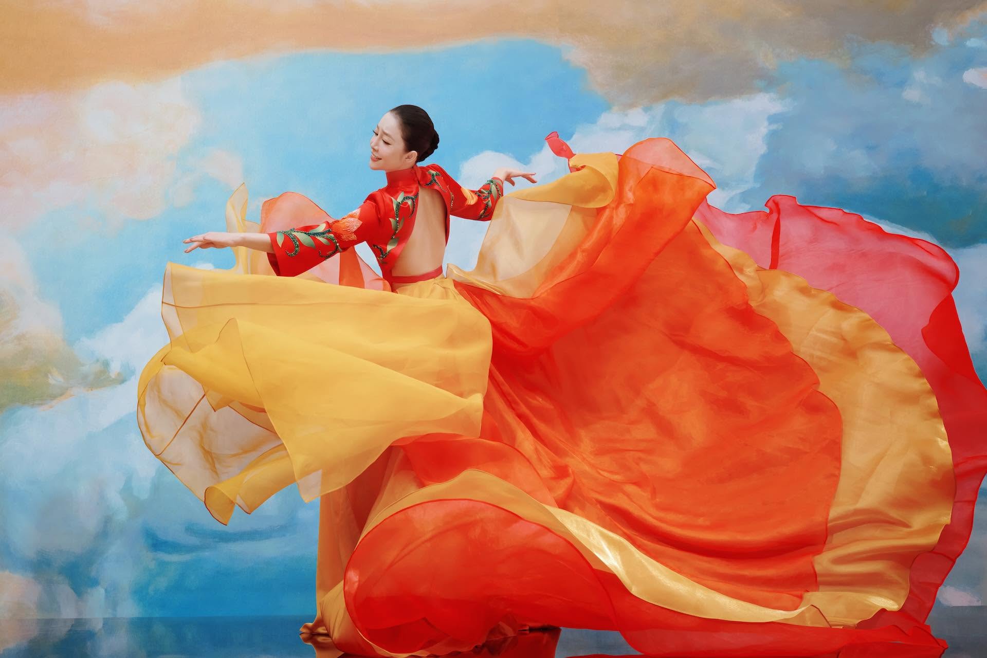 Linh Nga dancing in a flowing red, orange and yellow costume against a painted sky.