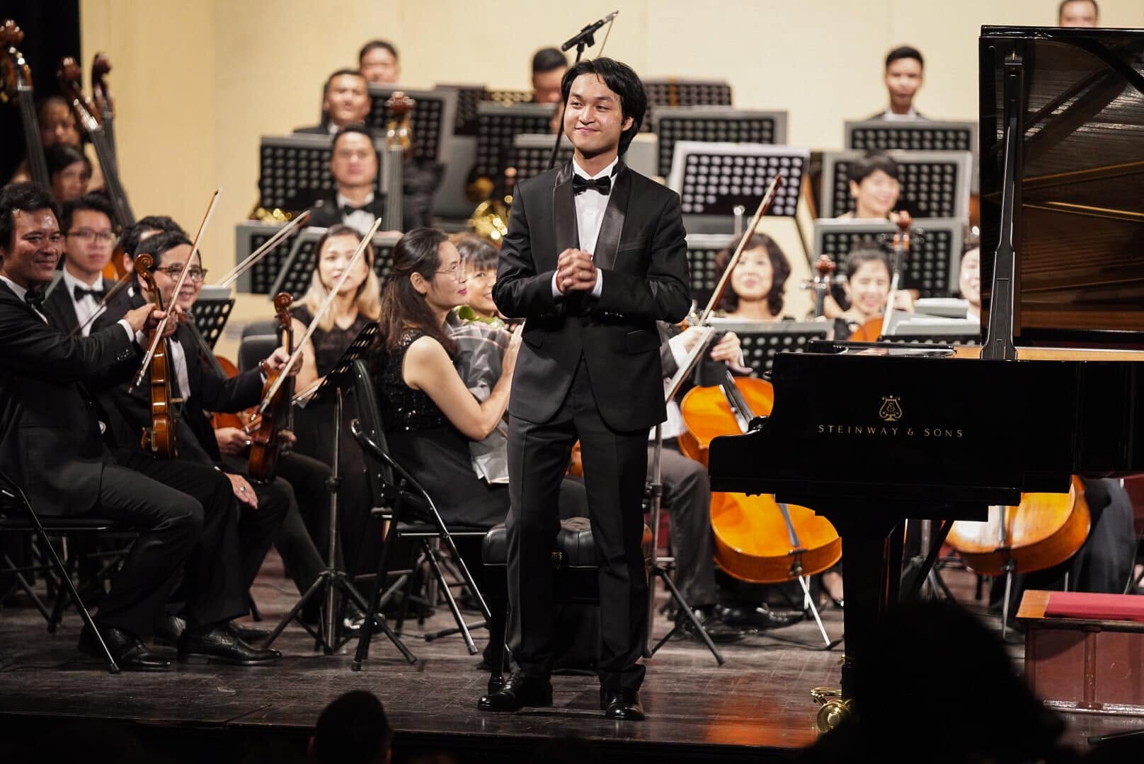 Nguyen Dang Quang in a tuxedo takes a bow beside a Steinway grand piano, the orchestra behind him.
